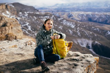 aktif genç kız sarı bir sırt çantası ile dağ kenarında oturur, dağ doğa ve güneş sahiptir