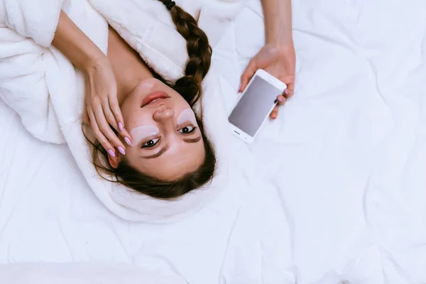 Jong meisje in witte robe ligt in bed, onder ogen patches, houdt de telefoon — Stockfoto