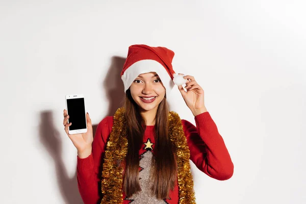 Glücklich lächelndes Mädchen mit roter Mütze auf dem Kopf, das ein Smartphone hält und auf das neue Jahr wartet — Stockfoto