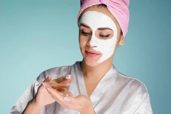 Carina ragazza con un asciugamano rosa sulla testa applicando una maschera idratante bianca sul suo viso — Foto Stock