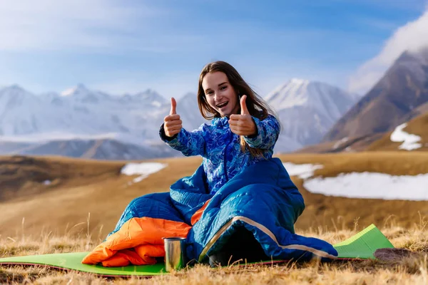 Mutlu genç kız mavi uyku tulumu başparmak gösterilmesini Kafkas Dağları'nın arka plan üzerinde oturan — Stok fotoğraf