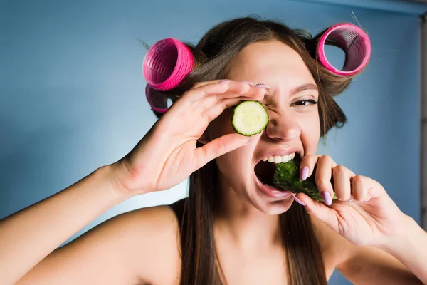 Funny mladá dívka držící okurky pro zvlhčení pokožky v obličeji, na hlavě natáčky — Stock fotografie