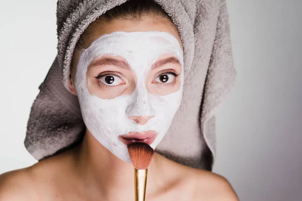 Όμορφη νεαρή κοπέλα με μια πετσέτα στο κεφάλι της εφαρμόζεται μια λευκή θρεπτική μάσκα στο πρόσωπό της με ένα πινέλο — Φωτογραφία Αρχείου