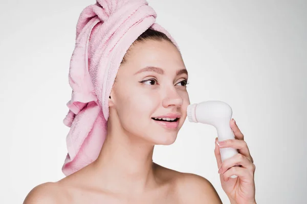 Una giovane ragazza carina con un asciugamano rosa sulla testa fa una pulizia profonda del codice del viso con una spazzola elettrica — Foto Stock