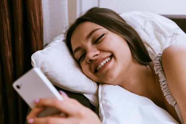 Glückliches junges Mädchen liegt im Bett, schaut ins Smartphone, lächelt — Stockfoto