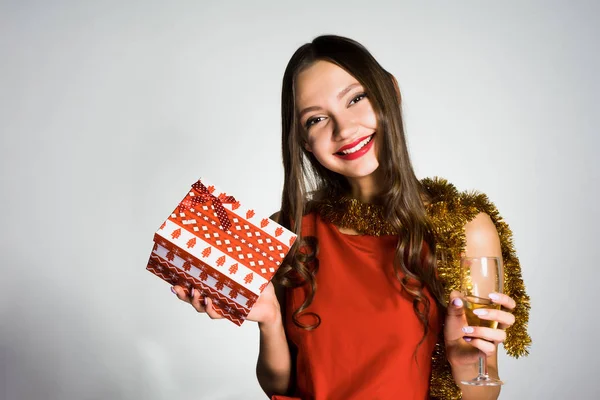 Счастливая улыбающаяся девушка в красном платье празднует Новый год, держит в руках подарок и шампанское — стоковое фото