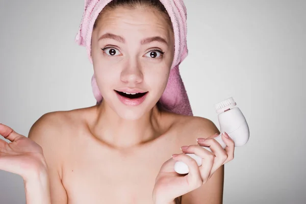 Jovem menina surpreso com uma toalha rosa na cabeça detém uma escova elétrica para limpar a pele do rosto — Fotografia de Stock
