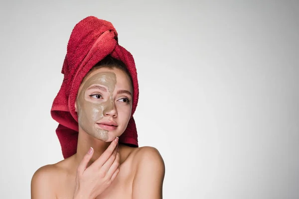 Roztomilá mladá dívka s červený ručník na hlavě použít jílová maska na polovinu tváře — Stock fotografie