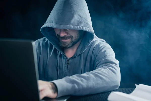 Hacker masculino em uma camisola com um capuz se senta atrás de um laptop — Fotografia de Stock