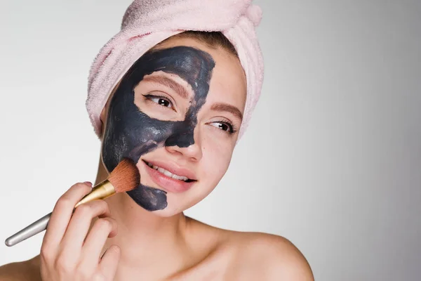 Μια γυναίκα με μια πετσέτα στο κεφάλι της μια καθαριστική μάσκα εφαρμόζεται στο πρόσωπό της σε ένα γκρίζο φόντο — Φωτογραφία Αρχείου