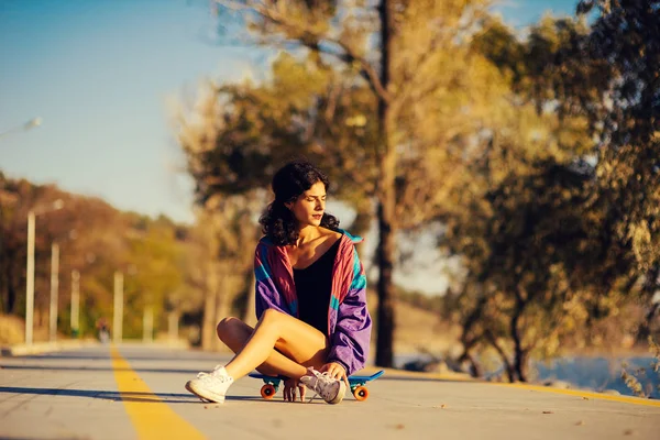 Beautiful woman is sitting on the skateboard on the background of the track — Stock Photo, Image