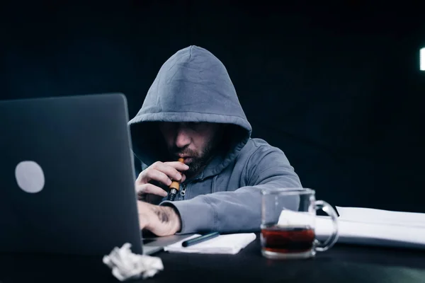 En mann som hacker i genser med hette, sitter bak en laptop og røyker en hanske. – stockfoto