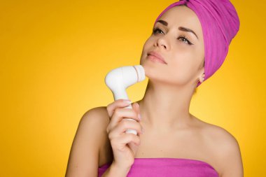 kadının kafasındaki bir havluyla yüzünü sarı bir arka plan üzerinde derin temizleme fırçası ile deri temizler