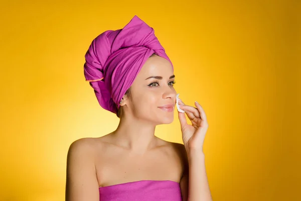 Kvinna med en handduk på huvudet på en gul bakgrund tar bort makeup med en bomull-skiva — Stockfoto