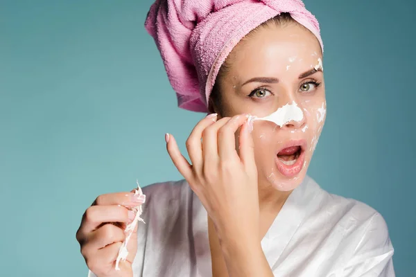 Женщина с полотенцем на голове снимает белую маску с лица — стоковое фото