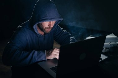 adam hacker'bir kazak bir kukuleta ile bir dizüstü bilgisayar arkasındaki odada oturur