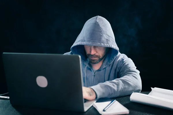 Um homem hacker está sentado em uma sala para um laptop fumar e algo imprime — Fotografia de Stock