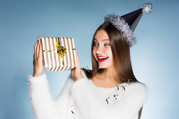 En glad kvinne i en festlig hatt som holder en gave på blå bakgrunn – stockfoto