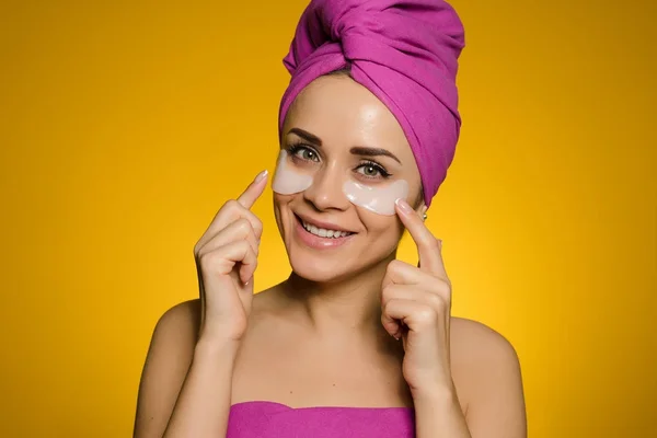 Ευτυχισμένη γυναίκα με μια πετσέτα στο κεφάλι της μπαλώματα κάτω από τα μάτια εφαρμόζεται σε κίτρινο φόντο — Φωτογραφία Αρχείου
