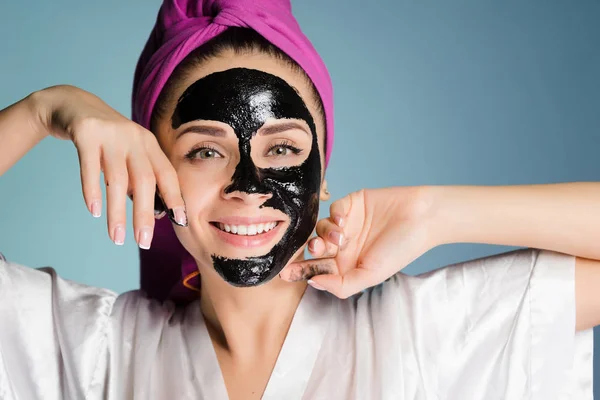Ευτυχισμένη γυναίκα με μια πετσέτα στο κεφάλι της μια καθαριστική μάσκα εφαρμόζεται πάνω στο δέρμα του προσώπου σε μπλε φόντο — Φωτογραφία Αρχείου