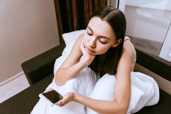 Trauriges Mädchen, das auf dem Bett sitzt und auf das Telefon schaut — Stockfoto