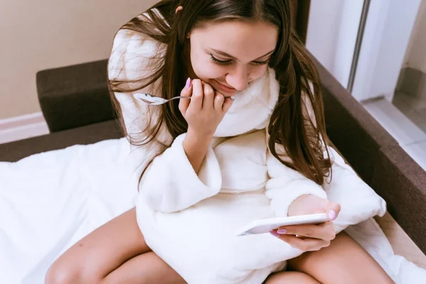 Szczęśliwa Kobieta w szlafroku siedzi na łóżku i patrzy na telefon — Zdjęcie stockowe
