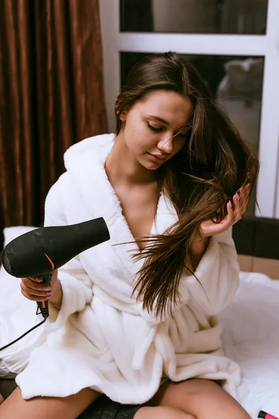 Vrouw met lang haar op het bed zit en droogt haar hoofd — Stockfoto