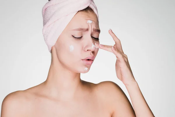 Женщина с полотенцем на голове наносит крем на кожу лица на сером фоне — стоковое фото