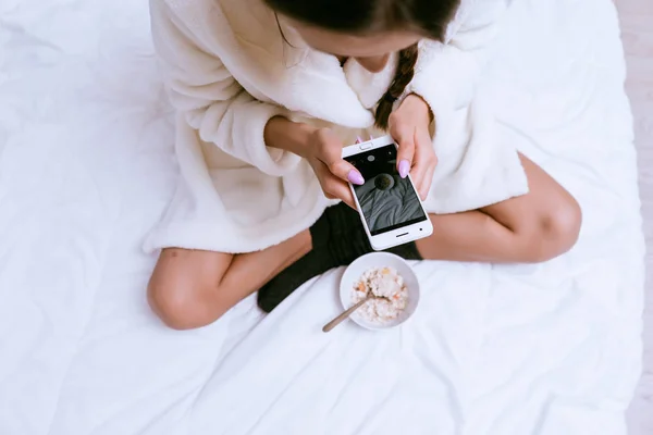 Женщина сидит на кровати и смотрит в телефон — стоковое фото