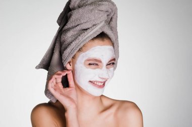 Kafasındaki bir havlu ile mutlu kadın gri bir arka plan üzerinde yüz cildi temizlemek için bir maske uygulanan
