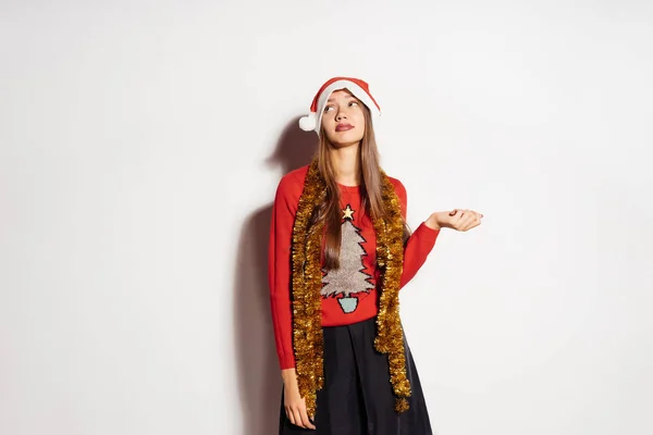 Divertida joven con una gorra roja de Año Nuevo y un suéter esperando un nuevo año y Navidad, en el cuello un oropel de oro —  Fotos de Stock