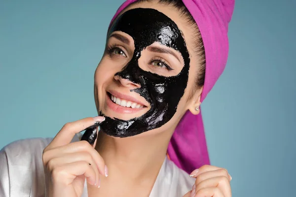 En leende flicka med en rosa handduk på huvudet tillämpas en svart mask för ansiktet — Stockfoto