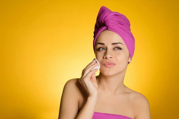 Menina bastante confiante com uma toalha rosa na cabeça limpa a pele do rosto com um disco de algodão — Fotografia de Stock