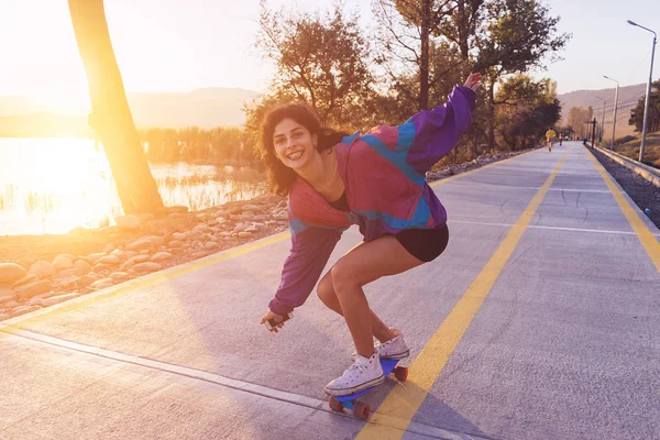 Ragazza felice in una giacca alla moda sta cavalcando su un longboard al tramonto — Foto Stock