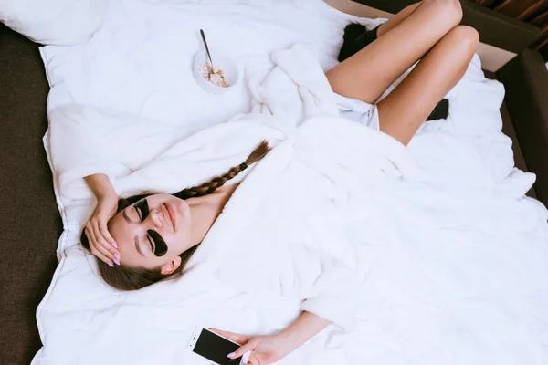 A cute laughing girl in a white robe lies on a bed, under the eyes of black patches — Stock Photo, Image