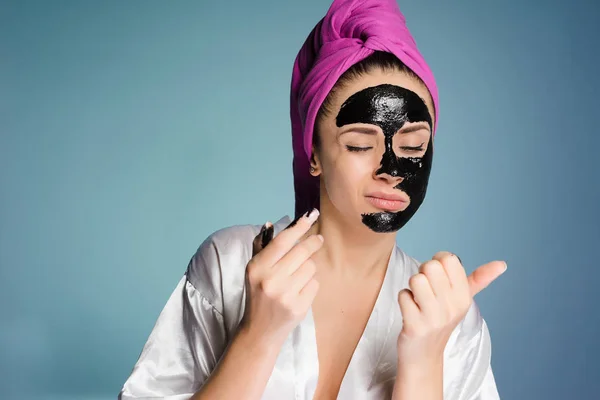 Linda joven con una toalla rosa en la cabeza pone en su cara una máscara negra contra el acné — Foto de Stock