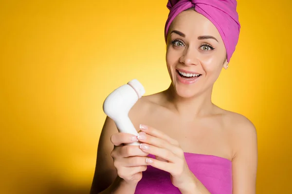 Felice ragazza con un asciugamano rosa sulla testa tiene una spazzola elettrica per pulire la pelle del suo viso — Foto Stock