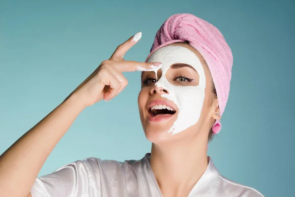 Divertida chica sonriente con una toalla rosa en la cabeza causando máscara blanca nutritiva en la cara —  Fotos de Stock