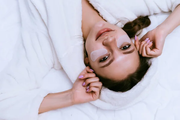 Joven chica atractiva en túnica blanca se encuentra en la cama, bajo los ojos manchas blancas —  Fotos de Stock