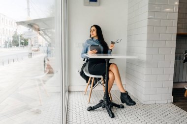 mavi gömlekli genç kız öğrenci bir kafede oturuyor, bir tablet tutar ve pencereden görünüyor