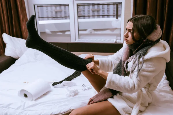 Θλιβερή νεαρή κοπέλα σε έναν λευκό terry ρόμπα βάζει στις ζεστές κάλτσες, αρρωσταίνει — Φωτογραφία Αρχείου