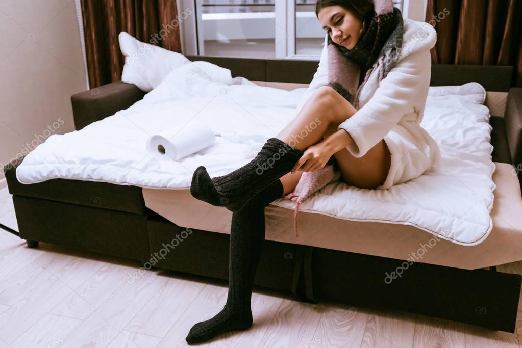 cute young girl in a white robe sits on the bed, puts on warm socks