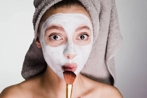 Une femme avec une serviette sur la tête après la douche a appliqué un masque sur la peau de son visage et tient une brosse près de son visage — Photo