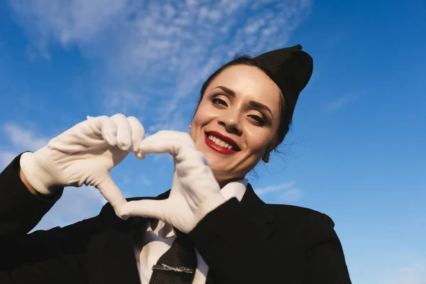 Счастливая стюардесса в форме на фоне голубого неба показывает сердце — стоковое фото