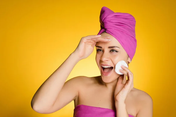 Donna felice con un asciugamano sulla testa rimuove il trucco con un disco di cotone su uno sfondo giallo — Foto Stock