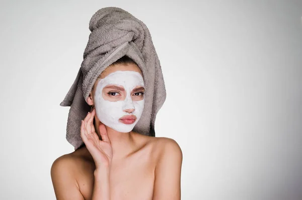 Γυναίκα με μια πετσέτα στο κεφάλι της εφαρμοστεί μια μάσκα στο πρόσωπό της σε ένα γκρίζο φόντο — Φωτογραφία Αρχείου