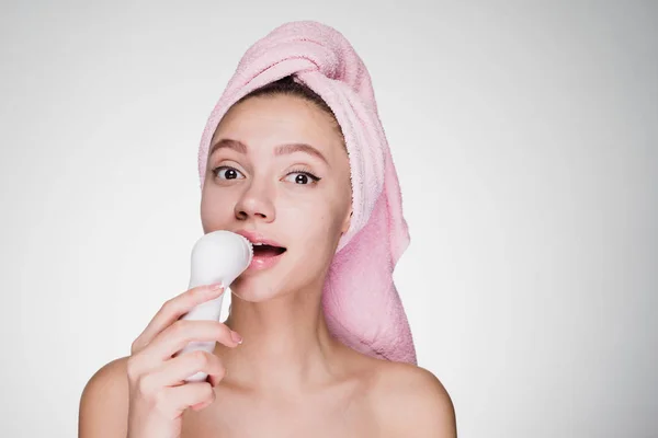 Una mujer pensativa con una toalla en la cabeza después de la ducha limpia su cara con un cepillo para una limpieza profunda —  Fotos de Stock