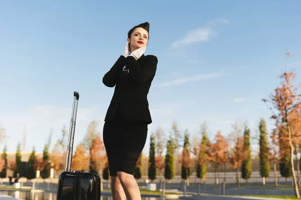 Nő a stewardess az egységes jelentő mellett a háttérégbolt bőrönd — Stock Fotó