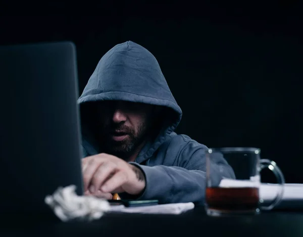 Pirate masculin dans un sweat-shirt avec une capuche se trouve derrière un ordinateur portable — Photo