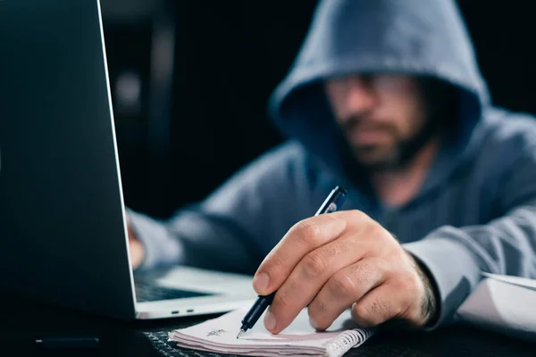 a man hacker in a sweatshirt with a hood sits behind a laptop and writes something in a notebook
